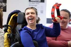 image shows a young girl in a wheelchair shaking a tambourine and smiling - taking part in the Inclusive Choir